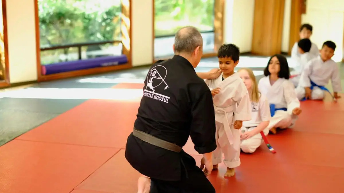 An instructor in a black uniform kneels on the red mats beside a row of young students in white uniforms at the academy