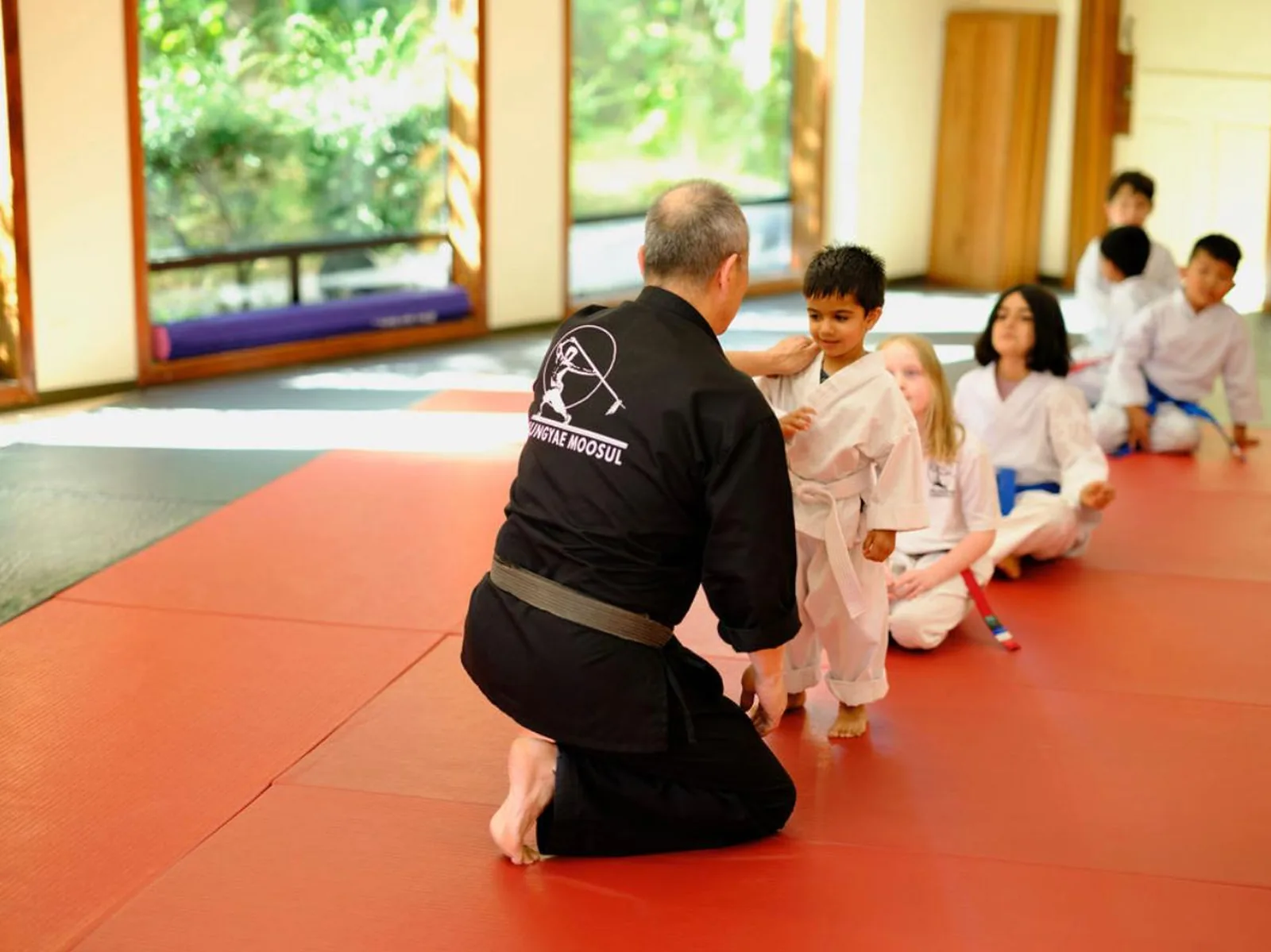 An instructor in a black uniform kneels on the red mats beside a row of young students in white uniforms at the academy