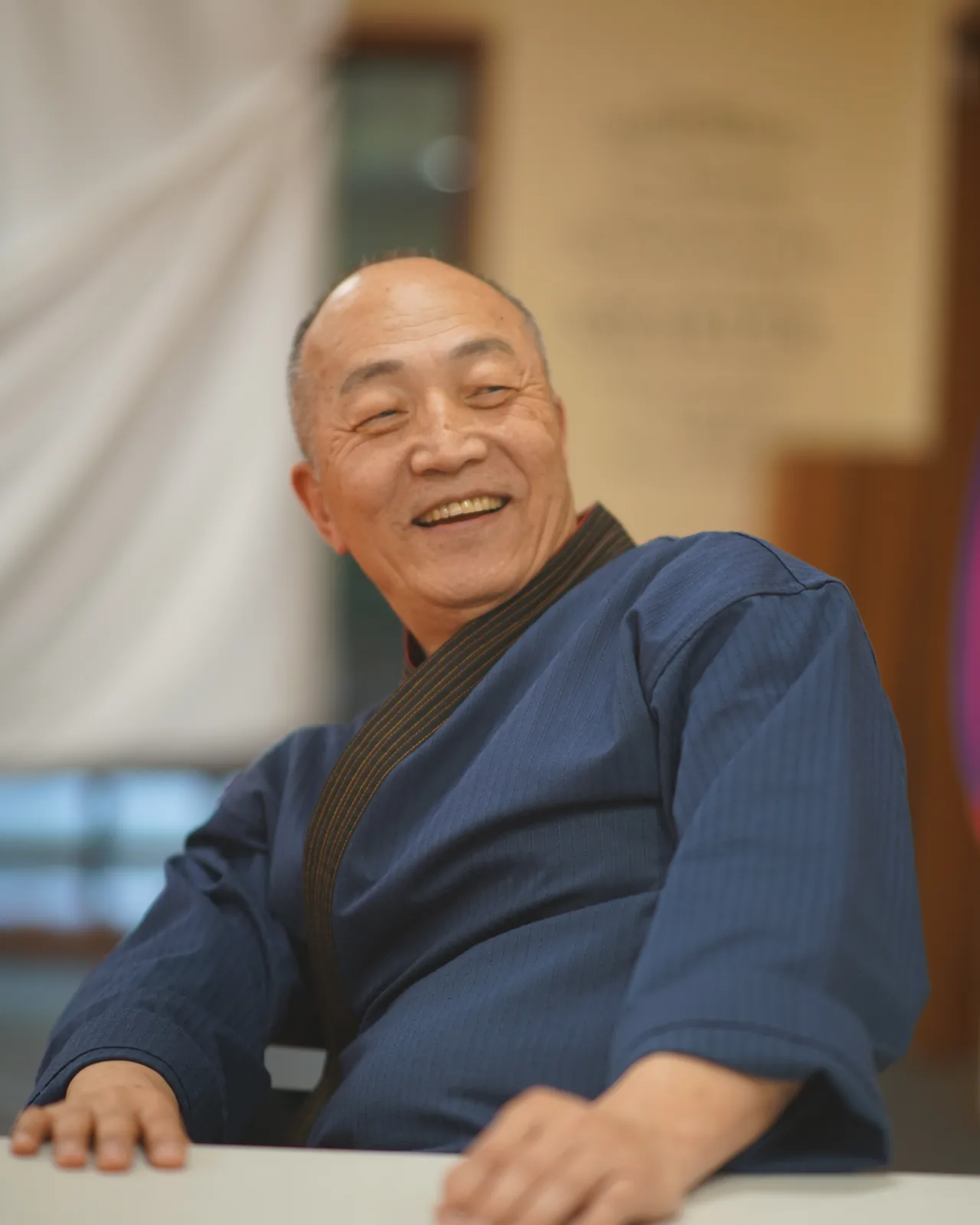 Grand Master Won Kuk Kim laughing as he leans on a table at the academy, wearing a navy dobok