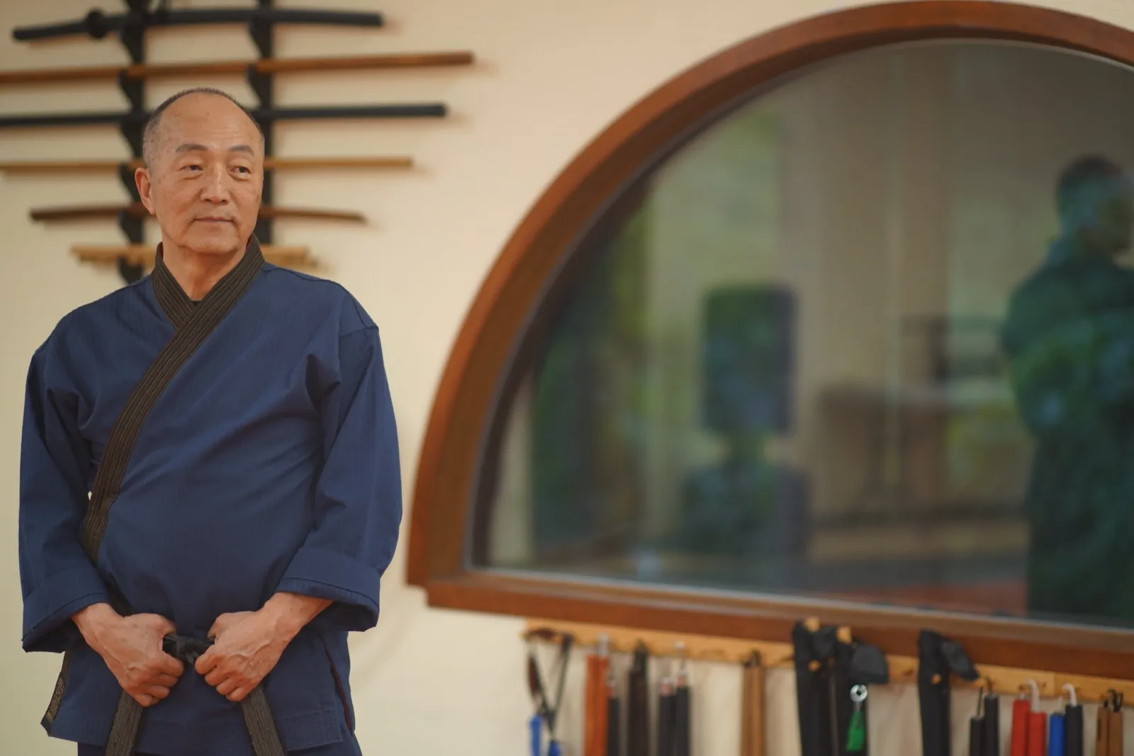 Grand Master Won Kuk Kim standing with his hands clasped beside an arched window and a weapon rack at the academy, wearing a navy dobok