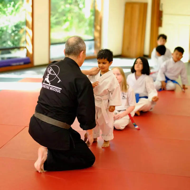 An instructor kneels on red mats beside a line of young students in white uniforms