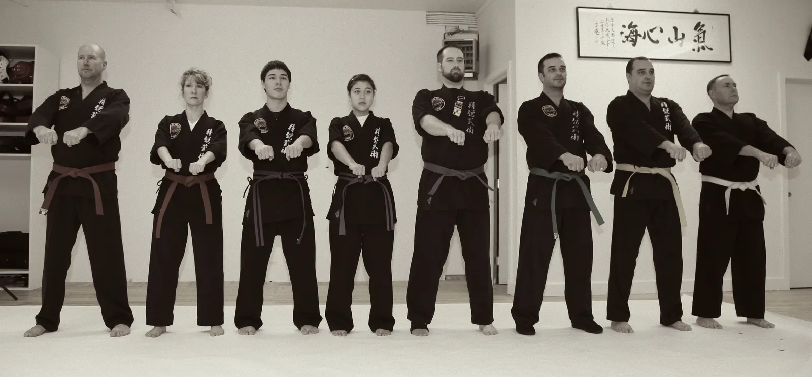 Eight adult students in black uniforms standing in a line with fists forward, calligraphy scroll behind them