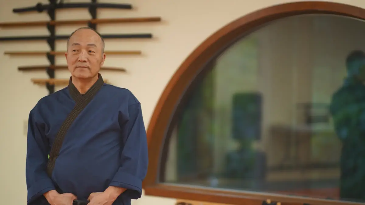 Grand Master Won Kuk Kim standing with hands clasped beside the arched window and weapon rack of the academy